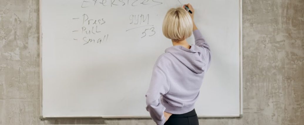 Female writing workout schedule on whiteboard in gym setting.